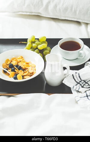 Breakfast in bed. Cornflakes with berries and milk, fruits and tea on a black tray. Stock Photo