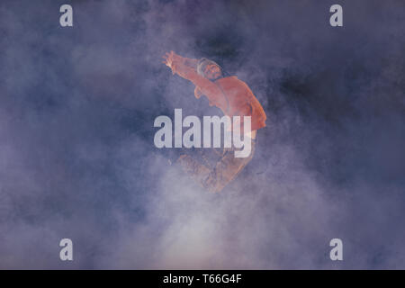 Male breakdancer jumps off high in the air while performing a cool dance move, a lot of smoke in the background. Stock Photo