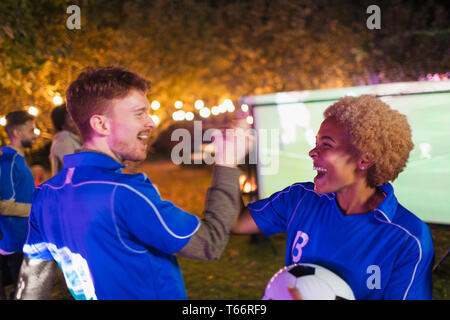 Happy friends cheering, watching soccer match on projection screen in backyard Stock Photo