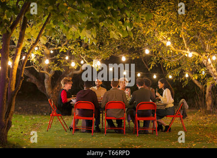 Friends enjoying dinner garden party under trees with fairy lights Stock Photo