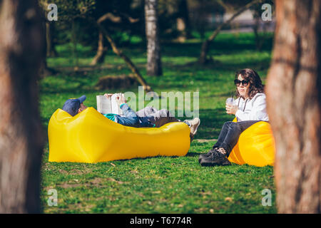 couple laying on yellow inflatable mattress in city park. reading book. drinking smoothies. leisure time Stock Photo
