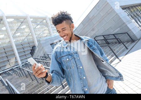 Young mulatto man standing near stairs on the city street browsing internet on smartphone smiling cheerful Stock Photo