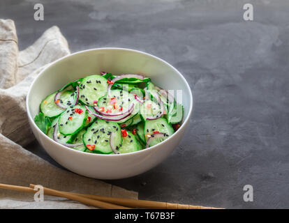 Asian cucumber salad with red onion, chilli pepper and black sesame in white bowl on gray concrete background. Stock Photo