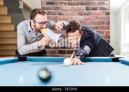 Billiard player aiming the ball while his friend disturbing him by using megaphone. Stock Photo