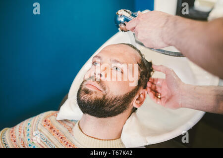 Barbershop theme. Barber is washing the head of the bearded man in the white sink with the black faucet in the barbershop. Stock Photo