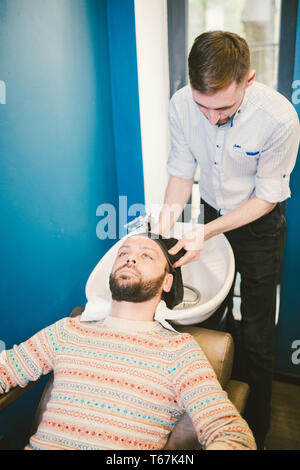 Barbershop theme. Barber is washing the head of the bearded man in the white sink with the black faucet in the barbershop. Stock Photo