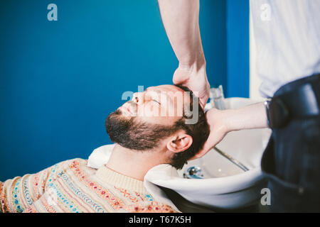 Barbershop theme. Barber is washing the head of the bearded man in the white sink with the black faucet in the barbershop. Stock Photo