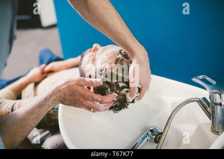 Barbershop theme. Barber is washing the head of the bearded man in the white sink with the black faucet in the barbershop. Stock Photo