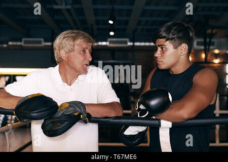 Young boxer listening to his mature trainer. Side view of a two sport person standing in boxer ring. Stock Photo