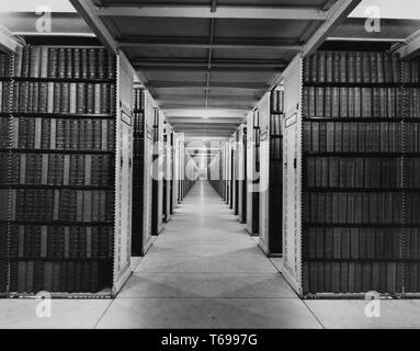 View of the center hallway of the stacks in a library.Black and white photograph of two slim, dark-haired young women, likely librarians, standing in profile, facing each other; the woman at left, who wears a band with a lamp around her head and a battery pack at her waist, holds a second head-lamp toward her colleague, whose hands are filled with a tray of slim books or papers; with shelves of hardcover books visible in the background, 1940. From the New York Public Library. () Stock Photo