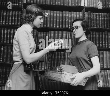 Black and white photograph of two slim, dark-haired young women, likely librarians, standing in profile, facing each other; the woman at left, who wears a band with a lamp around her head and a battery pack at her waist, offers a second head-lamp to her colleague, whose hands are filled with a tray of slim books or papers; with shelves of hardcover books visible in the background; located at the New York Public Library in New York, USA, 1948. From the New York Public Library. () Stock Photo