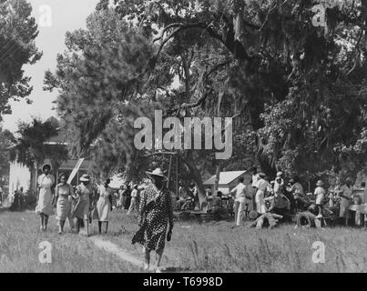 Black and white photograph of a group of African American men and women, enjoying themselves at a picnic or garden party; with a woman, wearing a large hat and a polka-dotted dress, walking toward the camera in the foreground, followed by a row of four young women wearing pale dresses, and with large trees and several buildings visible in the background; located in Beaufort, South Carolina, USA; photographed by Marion Post Wolcott, under the sponsorship of the United States' Farm Security Administration, July, 1939. From the New York Public Library. () Stock Photo