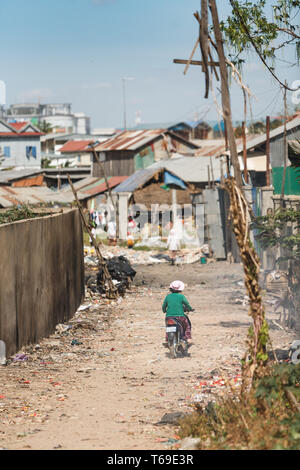 Woman in green sweater rides on motorbike through streets of garbage dump neighborhood in Phnom Penh Stock Photo