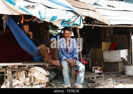 Two men are relaxing in the shade of a makeshift shelter. One man is sitting on the edge of the platform, while the other is reclining in a blue hammock. The shelter is constructed from a variety of materials, including corrugated metal, wood, and tarps. The men appear to be taking a break from their work or daily activities. Stock Photo