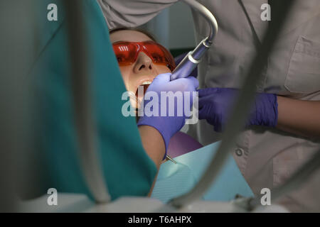Dentist is taking care about patient's tooth with black cavity on it using electric dental brush. Nurse with saliva ejector near the patient is helping to doctor. Woman's portrait in dental chair. Stock Photo