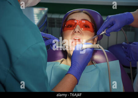Dentist is taking care about patient's tooth with black cavity on it using electric dental brush. Nurse with saliva ejector near the patient is helping to doctor. Woman's portrait in dental chair. Stock Photo