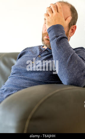 exhausted middle-aged man with a beard sits on sofa and rests his weary head in his hand Stock Photo
