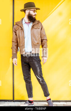 Full length portrait of a stylish bearded man dressed in jacket and hat on the bright yellow background outdoors Stock Photo
