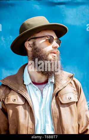 Close-up portrait of a stylish bearded man with hat and sunglasses on the blue wall background Stock Photo