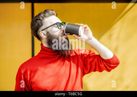 Portrait of a stylish bearded man in red swewater standing with coffee cup on the yellow background Stock Photo