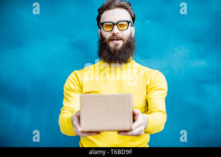 Stylish bearded man in yellow sweater standing with gift box on the blue background Stock Photo