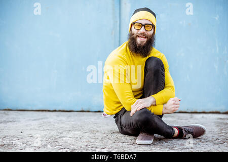 Portrait of a stylish man in yellow sweater sitting on the blue wall background outdoors Stock Photo