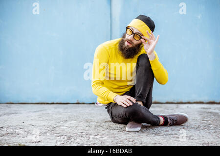 Portrait of a stylish man in yellow sweater sitting on the blue wall background outdoors Stock Photo