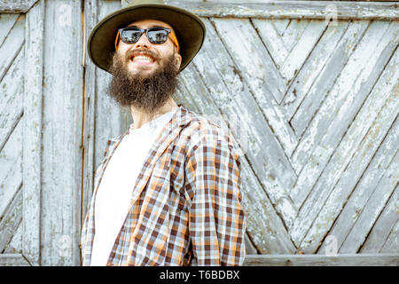 Portrait of a stylish positive man in checkered shirt and hat on the wooden gate background outdoors Stock Photo