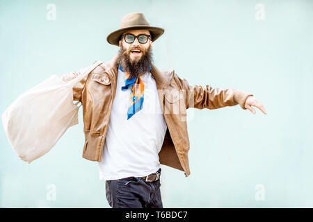 Portrait of a stylish man dressed in jacket, hat and scarf with bag on the turquoise background outdoors Stock Photo