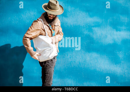 Portrait of a stylish bearded man dressed in jacket and hat standing with bag on the blue background outdoors Stock Photo