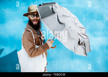 Portrait of a stylish bearded man dressed in jacket and hat standing with bag on the blue background outdoors Stock Photo