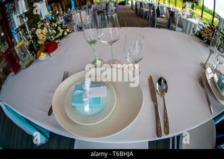 Table setting with handmade gift box on plate, glasses, forks, spoons and knives for wedding reception. Festive decorations and items for food, arrang Stock Photo