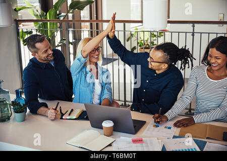 Diverse group of smiling young businesspeople high fiving together while working together at an office desk Stock Photo