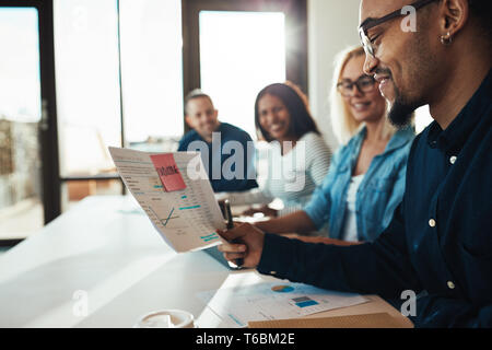 Smiling young African American office worker reading paperwork while sitting with a diverse group of colleagues in an office Stock Photo