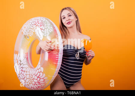 Attractive young woman in swimming suit holding a swim ring and fresh cocktail over yellow background. Ready for summer vacation. Beatiful blonde. Relaxing time. Stock Photo
