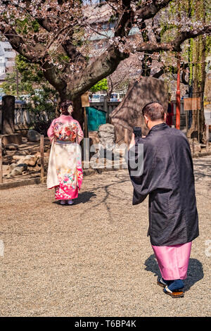 25 March 2019: Tokyo, Japan - Man photographing woman under cherry blossom in the grounds of the Asakusa Buddhist shrine in Tokyo; both are wearing... Stock Photo