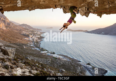 Female rock climber hanging upside down on challenging route in cave at sunset, resting before keeping on her attempt Stock Photo