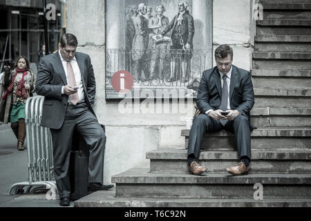 Two men in suits are taking a break to text and phone on the stairs of the old Stock Exchange as they are leaving the NYSE Euronext Stock Exchange on Wall Street. Stock Photo