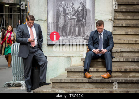 Two men in suits are taking a break to text and phone on the stairs of the old Stock Exchange as they are leaving the NYSE Euronext Stock Exchange on Wall Street. Stock Photo