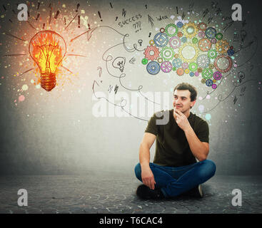 Cheerful man sitting on the floor smiling looking at a bright light bulb on the wall. Idea concept, positive thinking as colorful gear brain above hea Stock Photo