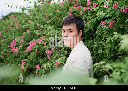 Portrait of serious handsome young man standing over green tree background, summer in the city Stock Photo