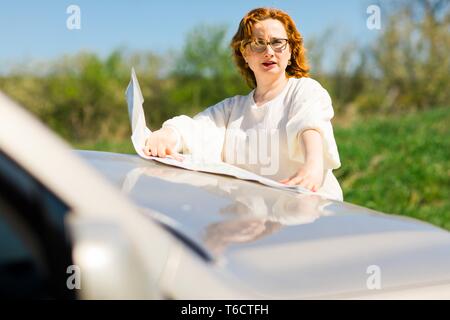 Attractive woman checking position in paper map on bonnet of the car - lost on trip in nature Stock Photo