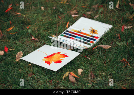 Watercolor painting of yellow red autumn maple leaf on white paper and box of used paints lying on green grass among fall leaves. Still life of hobby  Stock Photo