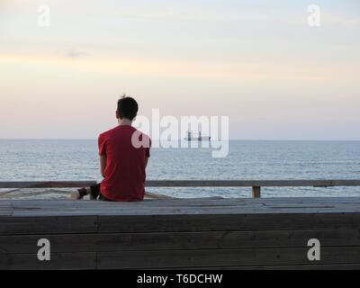 Young man sitting alone at the promenade looking at ship anchored off the coast, listening to music, ( concept of loneliness, relaxation). Stock Photo