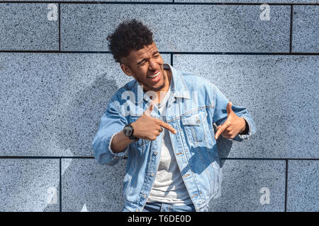 Young african american man standing isolated on concrete wall pointing down looking aside laughing cheerful close-up Stock Photo