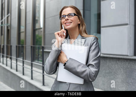 Young businesswoman wearing eyeglasses standing on the city street holding notebook and pen looking up thinking of creative ideas smiling dreamful Stock Photo