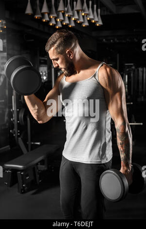 Bearded man bodybuilder standing at gym holding dumbbells looking at biceps curious right technique Stock Photo