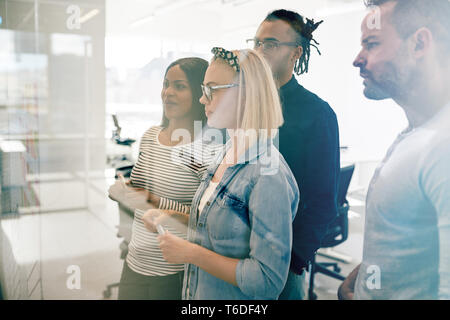 Diverse group of young work colleagues using sticky notes to brainstorm together on the glass wall of a modern office Stock Photo
