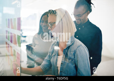 Smiling young businesswoman using sticky notes to brainstorm with her diverse team on the glass wall of a modern office Stock Photo