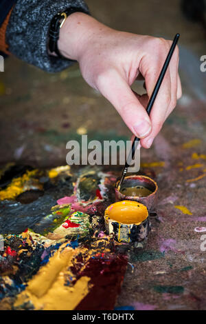 High angle close up of human hand dipping paintbrush into small pot of yellow oil paint. Stock Photo